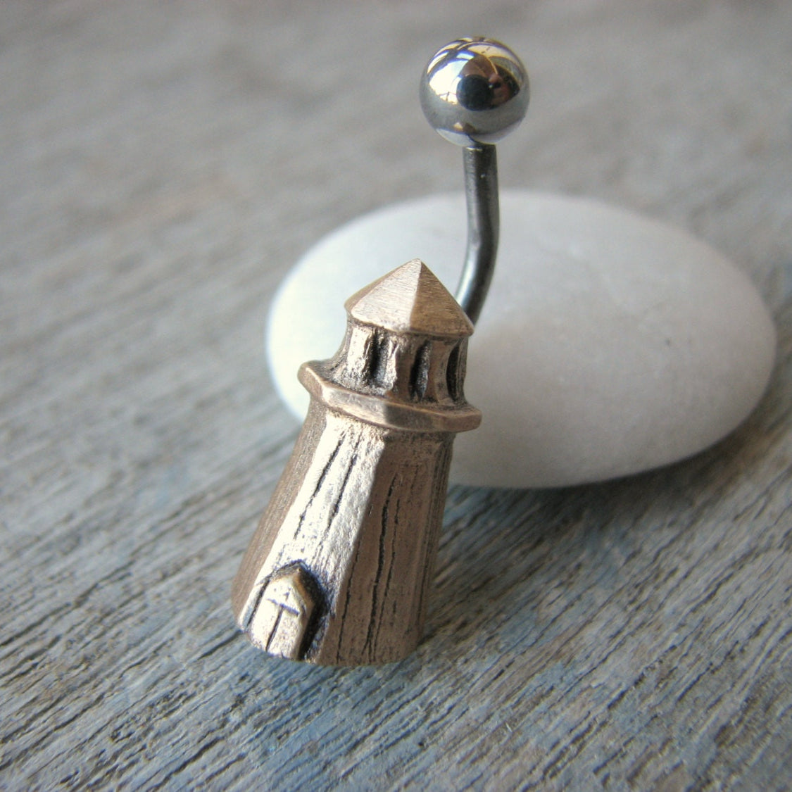 A belly button ring with a lighthouse charm on a white stone, the ring has a bronze lighthouse attached to a curved barbell.