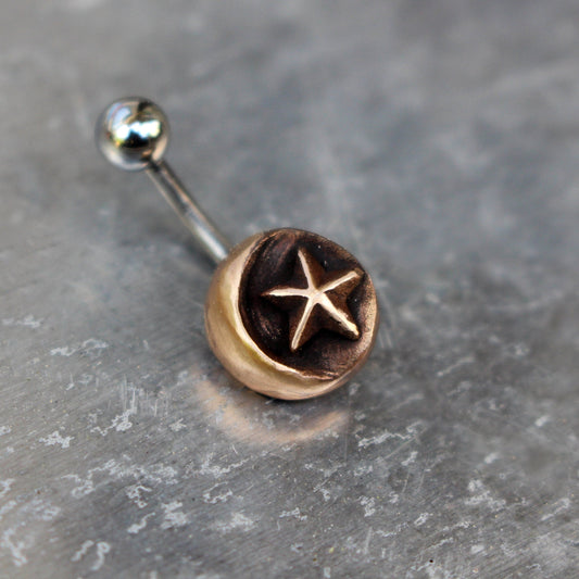 A celestial belly button ring with a crescent moon and a star made in solid bronze attached to one end of a curved barbell, with the other end being a ball end.