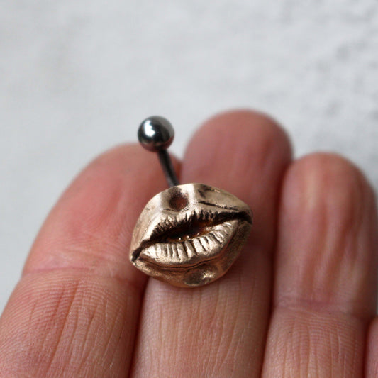 A belly button ring with a bronze mouth and lips design, held between fingers.