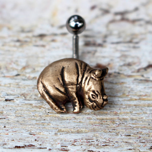 A belly button ring with a bronze rhinoceros charm attached to one end, set against a wooden background.