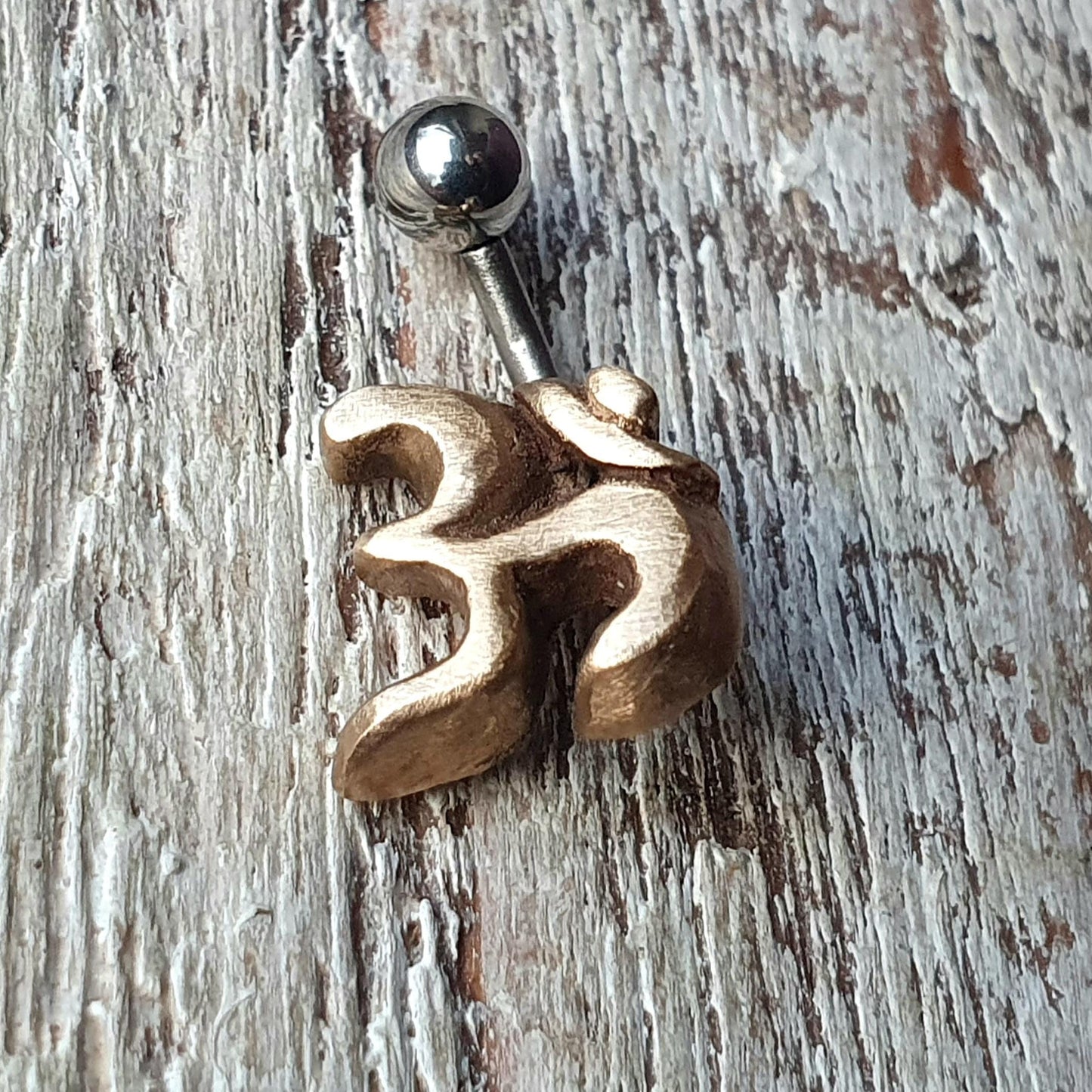 A belly button ring with a bronze 3D ohm symbol on one end and a screw on steel retaining ball on the other, placed against a wooden background.