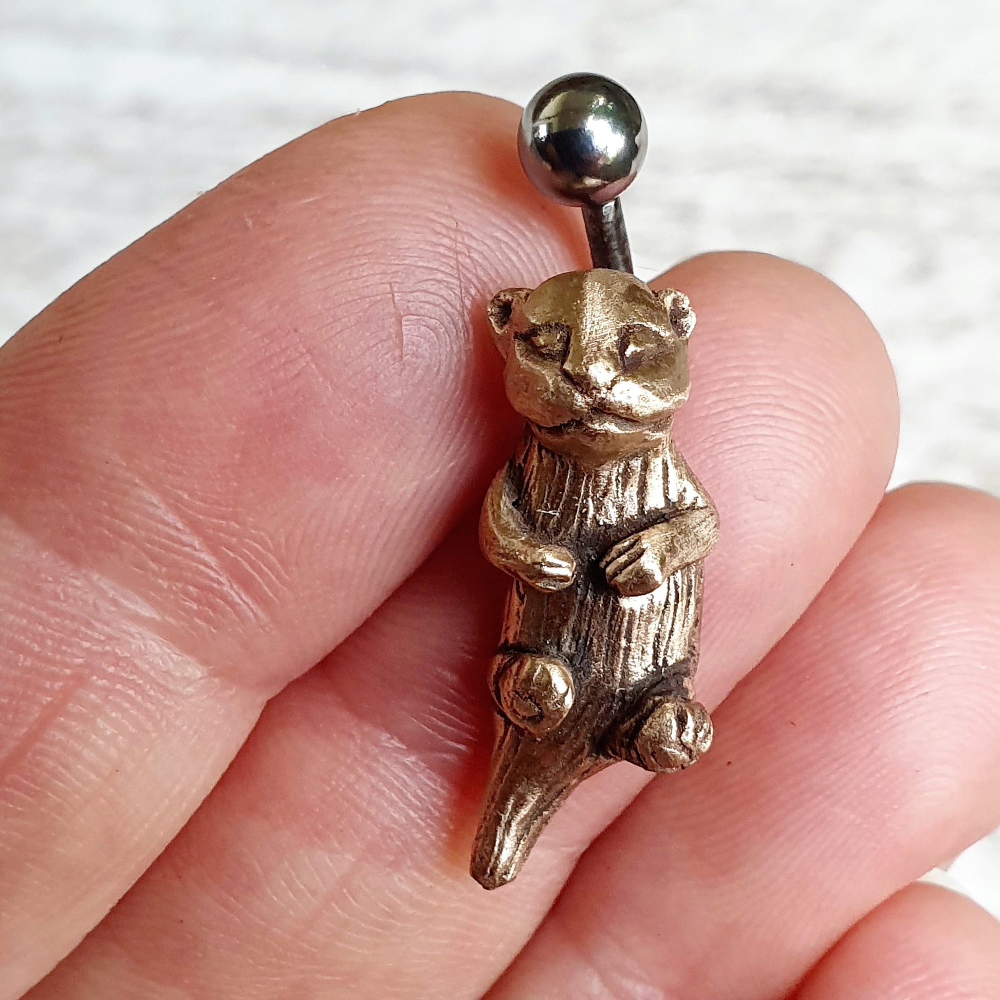 A sea otter belly button ring with a bronze sculptural otter on a curved barbell, held between fingers.