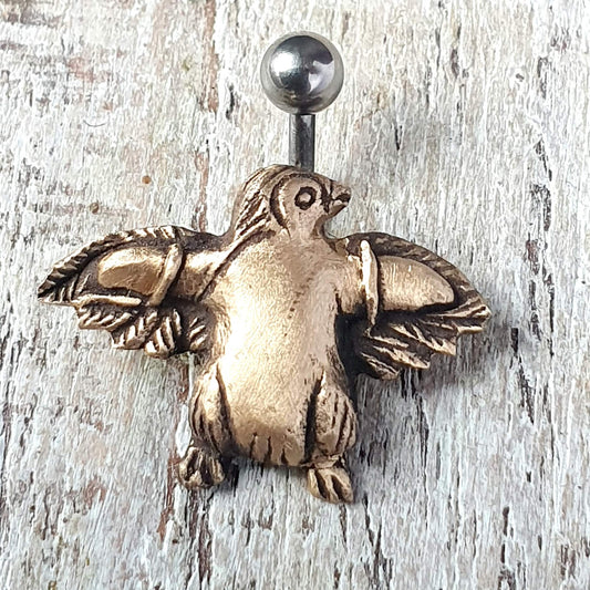 A bronze-colored belly button ring with a 3D sculpted design of a flying penguin attached to a barbell, set against a wooden background.