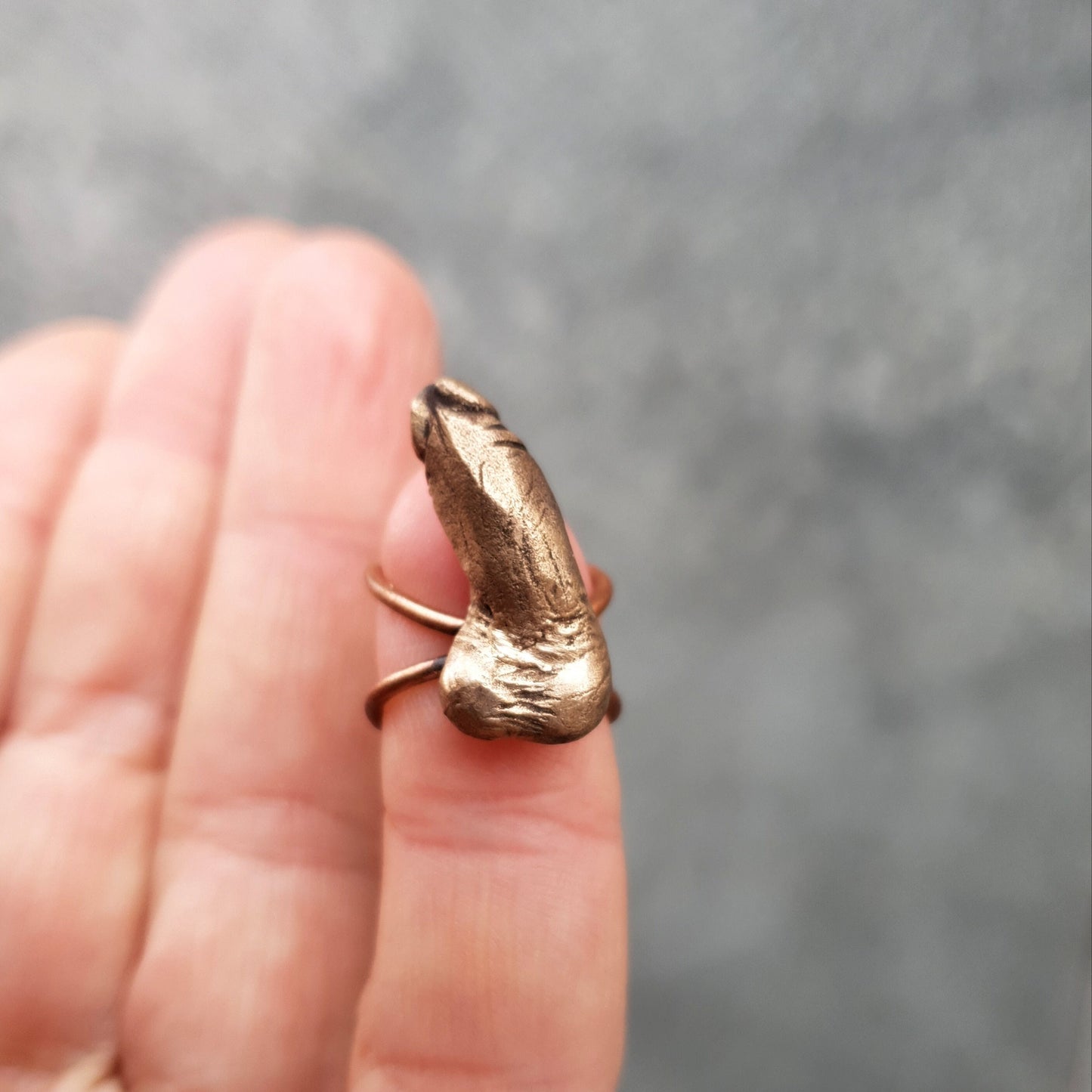 A bronze ear cuff in the shape of a miniature erected penis, displayed on a person's finger.