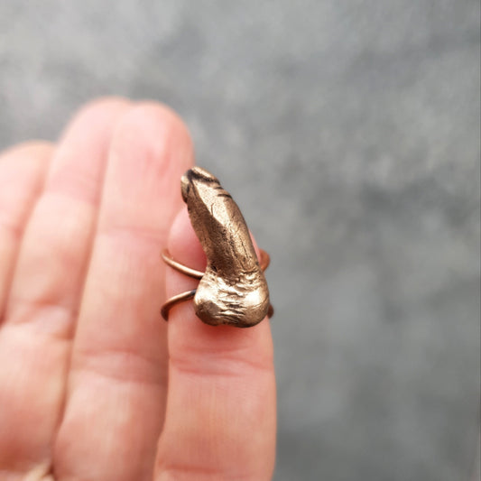 A bronze ear cuff in the shape of a miniature erected penis, displayed on a person's finger.