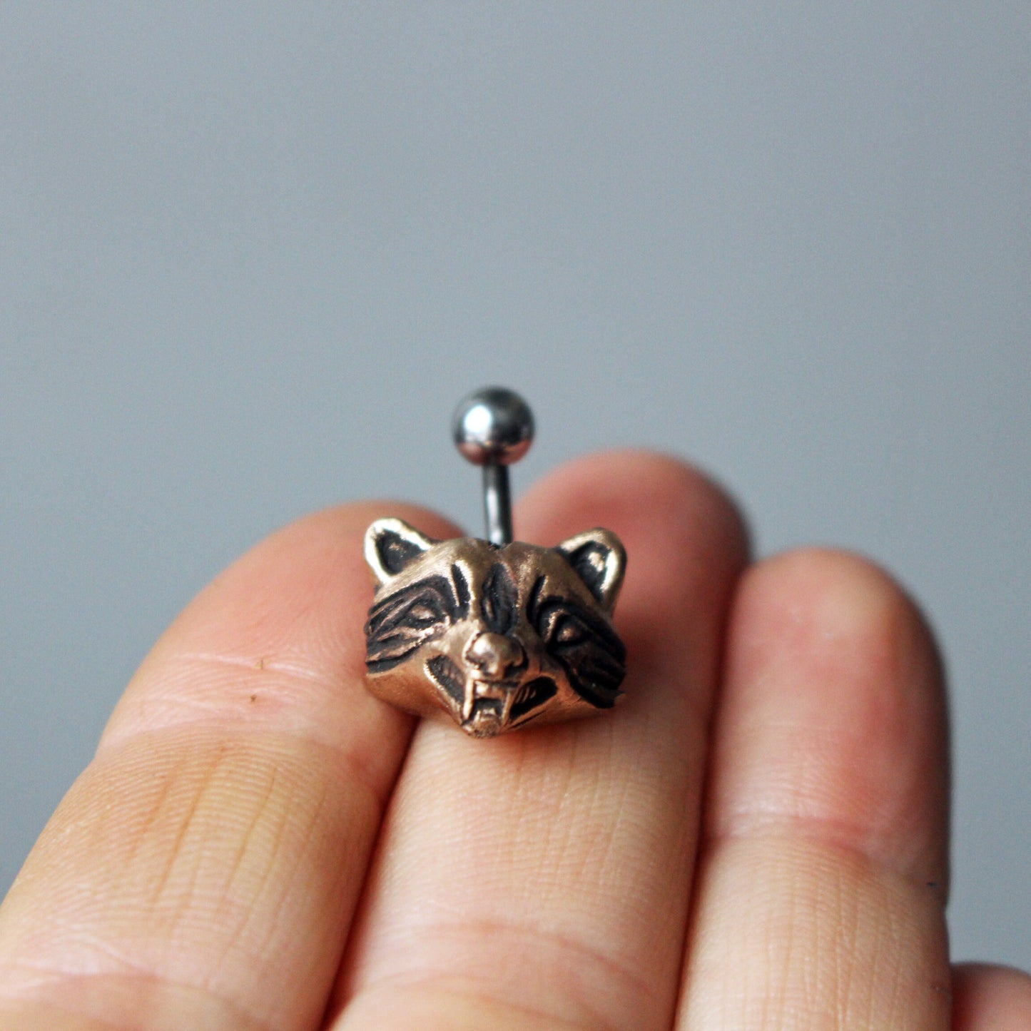 A bronze and titanium belly button ring with a raccoon head design held between fingers.