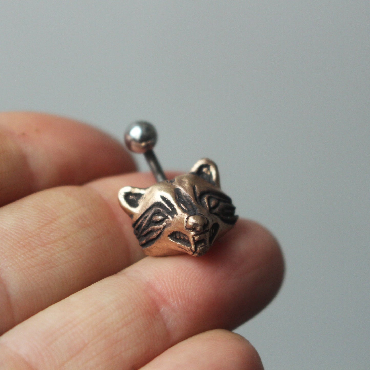 A bronze and titanium belly button ring with a raccoon head design held between fingers.