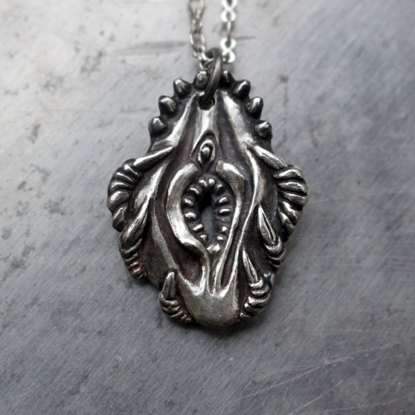 A silver-colored pendant featuring a detailed design of vagina dentata, intended to be worn as a necklace.