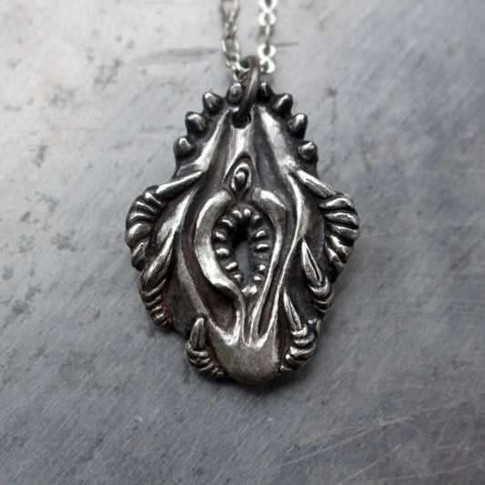 A silver-colored pendant featuring a detailed design of vagina dentata, intended to be worn as a necklace.