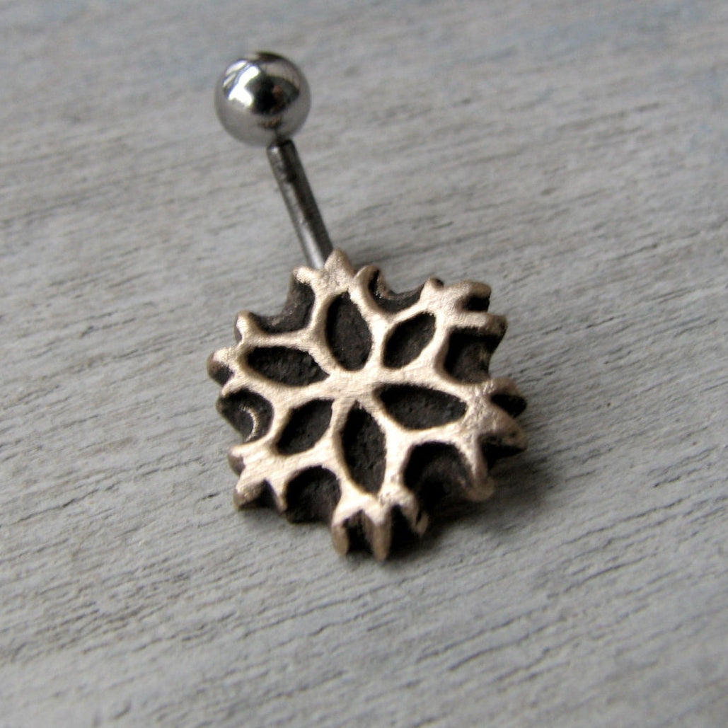 A snowflake-shaped belly button ring with a curved barbell, made of either titanium or surgical steel, displayed against a wooden background.