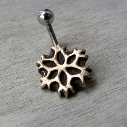 A snowflake-shaped belly button ring with a curved barbell, made of either titanium or surgical steel, displayed against a wooden background.