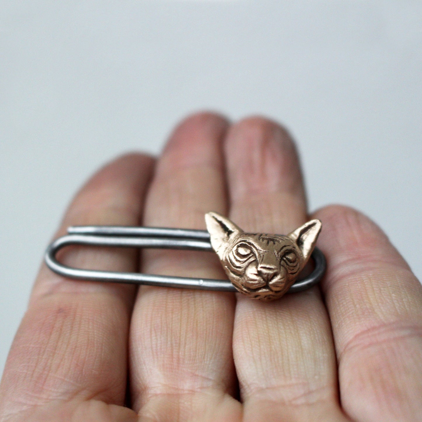 A bronze-colored hairless sphynx cat tie clip forged from stainless steel wire being held in a person's hand.
