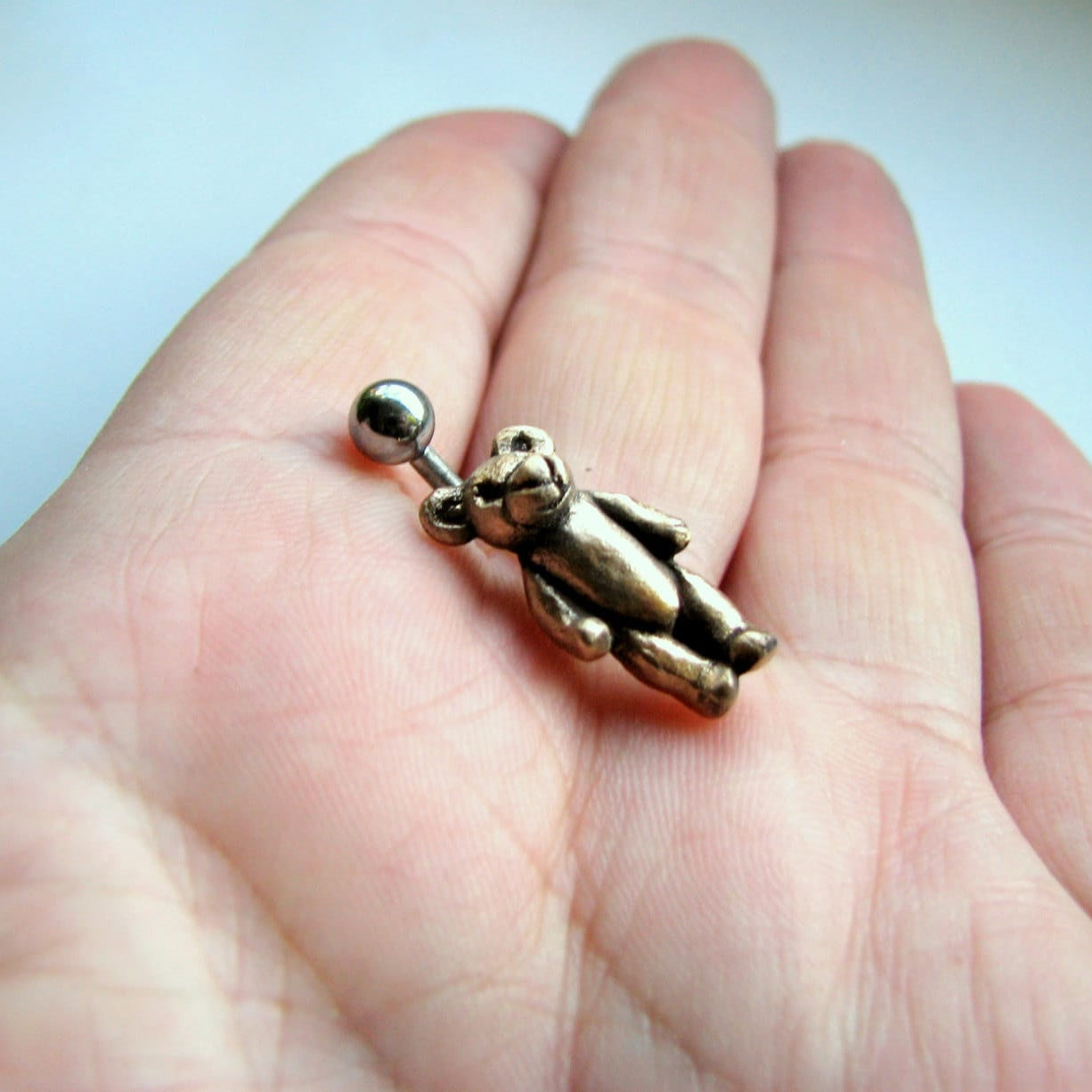 A bronze belly button ring in the shape of a teddy bear, held between fingers.