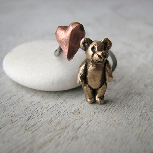 A bronze teddy bear ring with a pink copper heart on a white stone, against a wooden background.