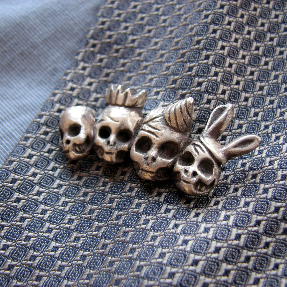 A pin brooch featuring a four skulls wearing partyhats, such as a crown and bunny ears, on a metallic surface.