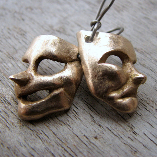 A pair of bronze theater mask earrings, one depicting a comedy mask and the other a tragedy mask, placed on a wooden surface.