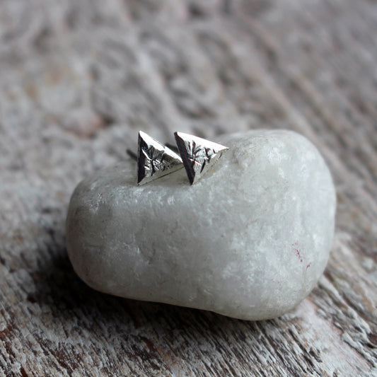 A pair of silver triangle post earrings with an uneven rough surface placed on a white pebble, with the earrings sparkling slightly in the light.