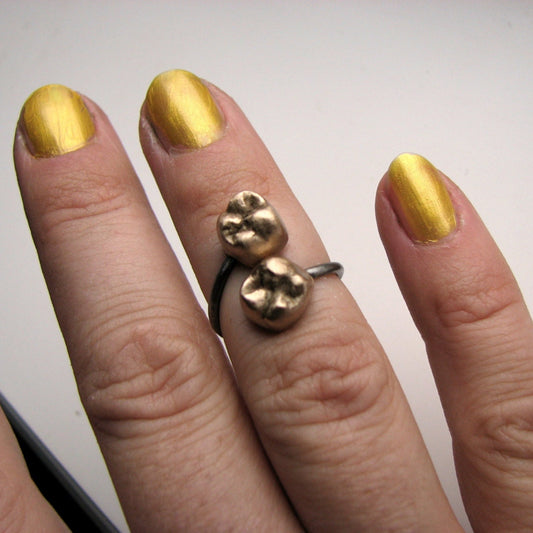 A hand with gold painted nails wearing an adjustable ring featuring a bronze-colored tooth design.