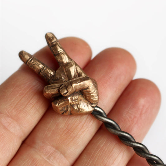 A hand holding a bronze hair stick with a sculpted miniature hand making a peace sign.