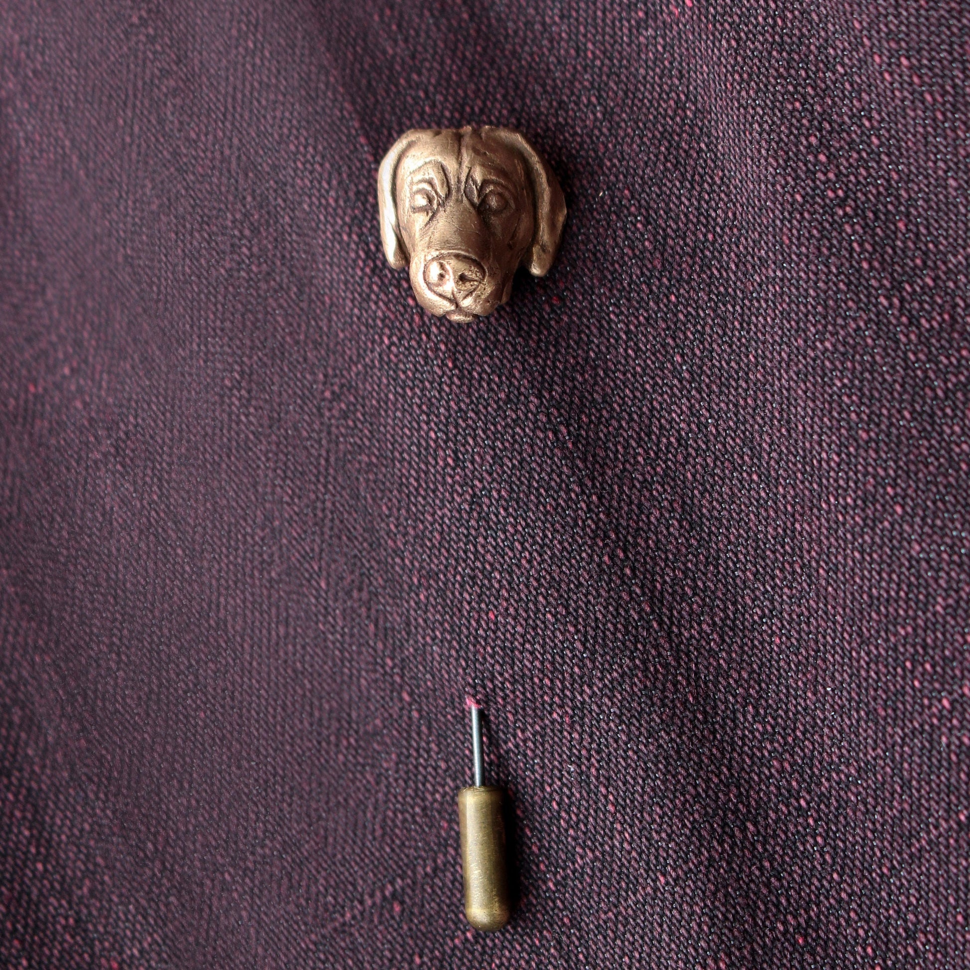 A bronze weimaraner dog head lapel stick pin made of bronze and stainless steel, on a purple fabric surface.
