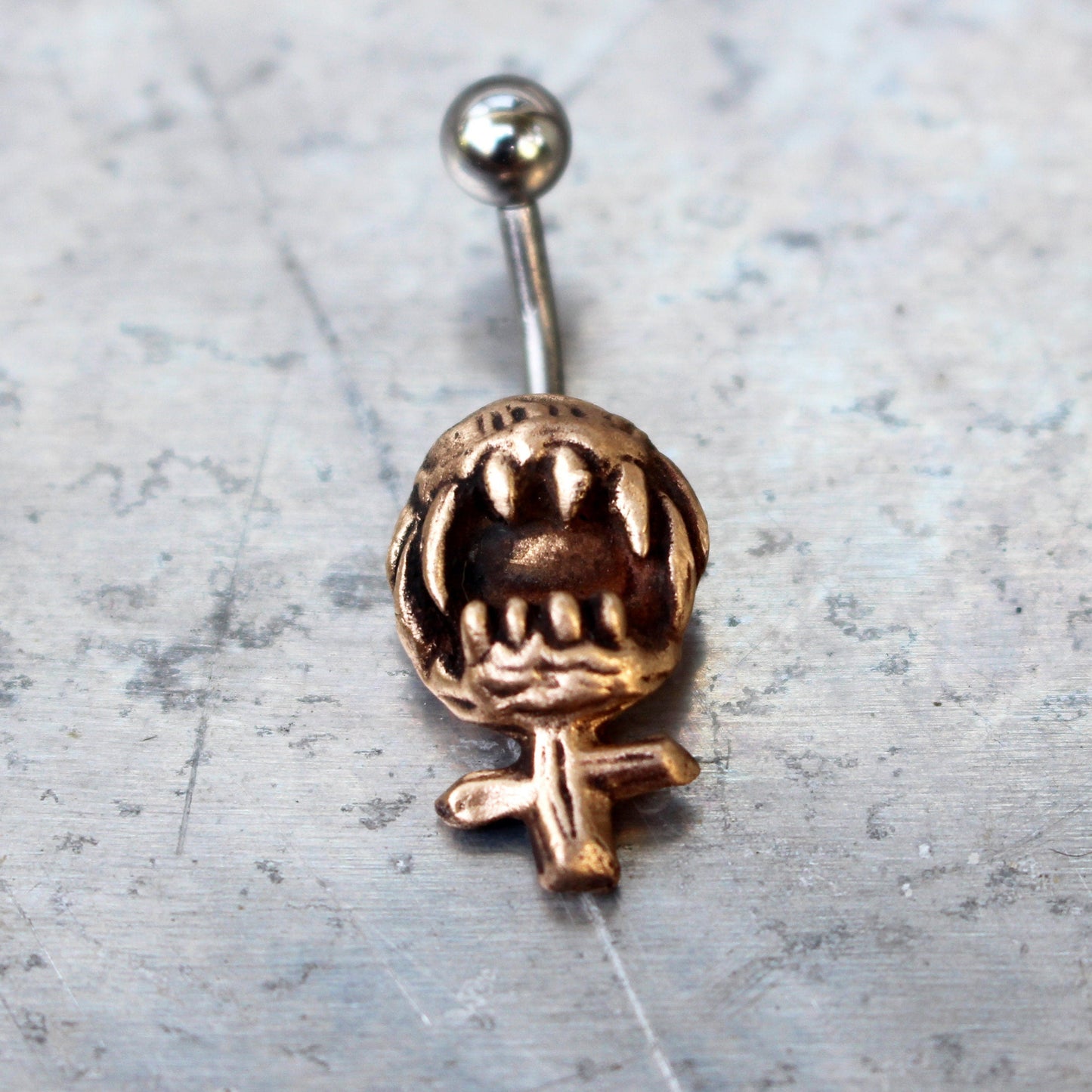 A belly button ring with a bronze feminist symbol design, featuring a barbell with either titanium or surgical steel material.