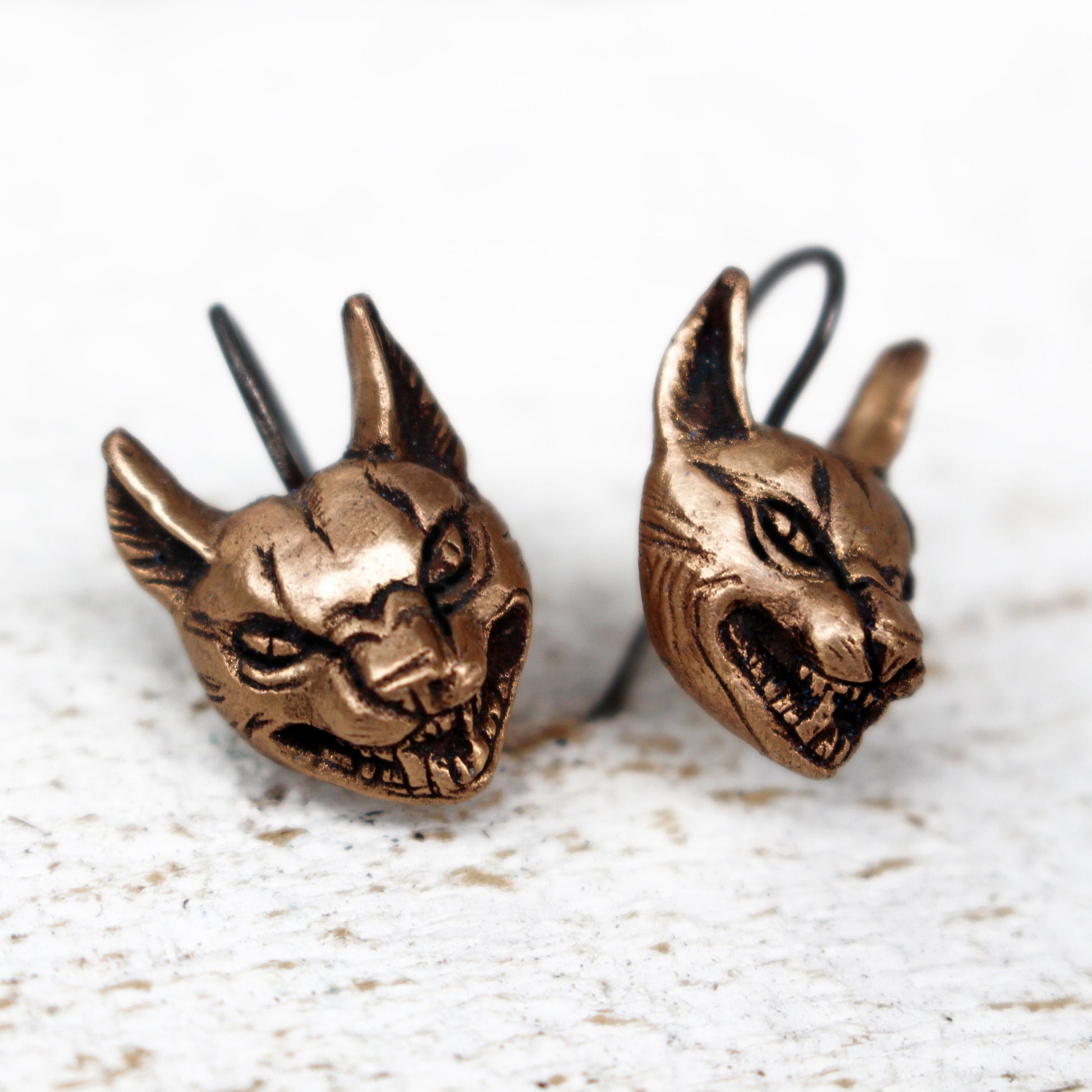 A pair of bronze dangle earrings in the shape of wild cat heads, displayed against a white background.