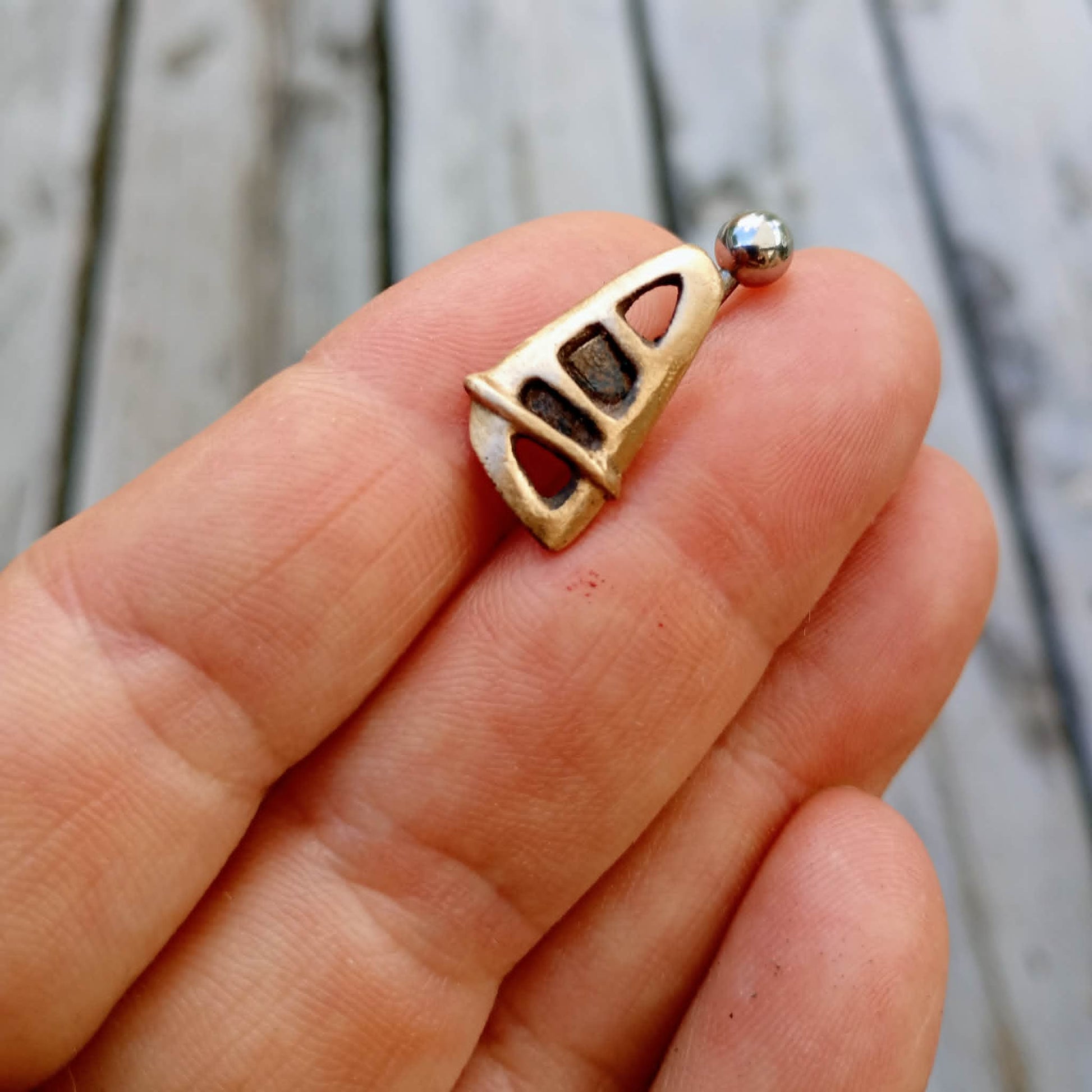 Small bronze and titanium windsurfing sain belly jewelry held between fingers against a wooden background