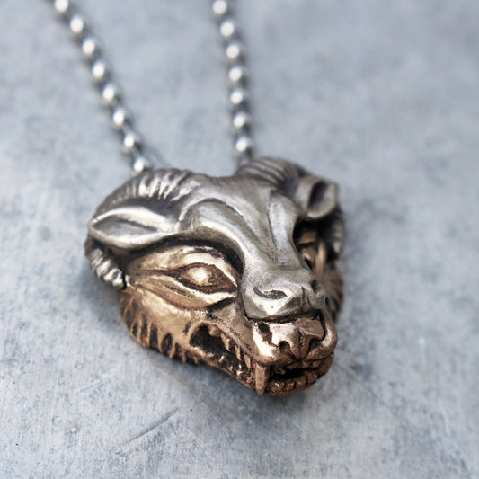 A pendant necklace with a wolf's head sculpted in yellow bronze with a sheep's mask in a contrasting white bronze, displayed on a chain.